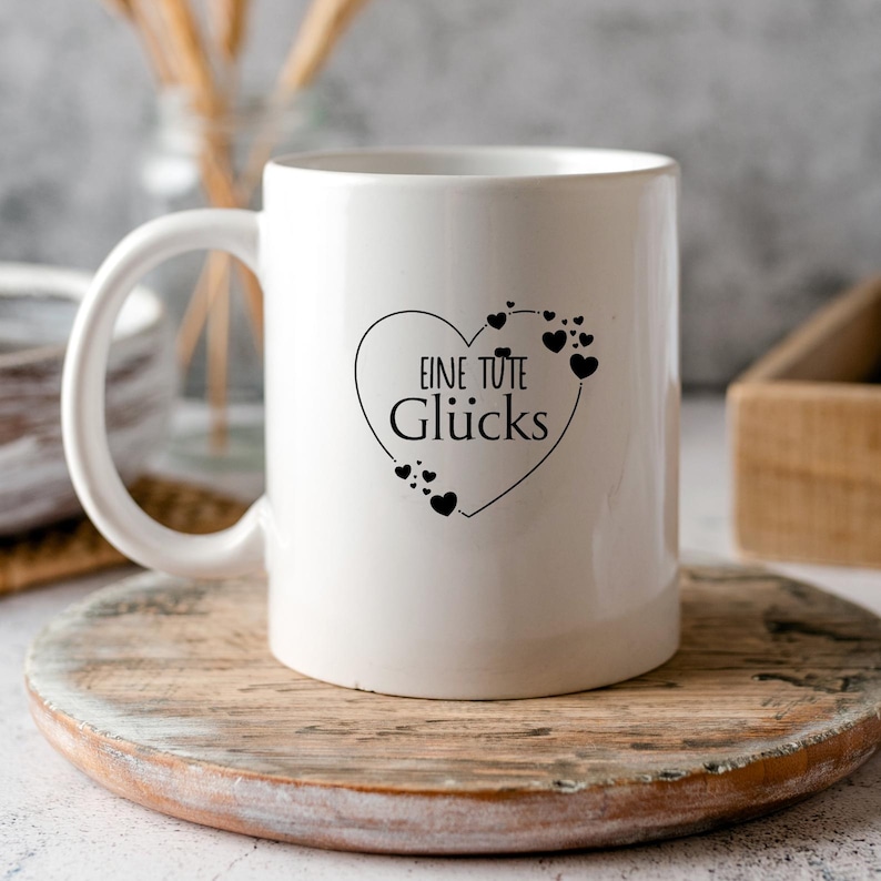 K&ouml;nnte beinhalten: Wei&szlig;e Keramiktasse mit einem schwarzen Herzdesign und dem Text "Eine Tute Gl&uuml;cks". Die Tasse steht auf einem Untersetzer aus Holz.