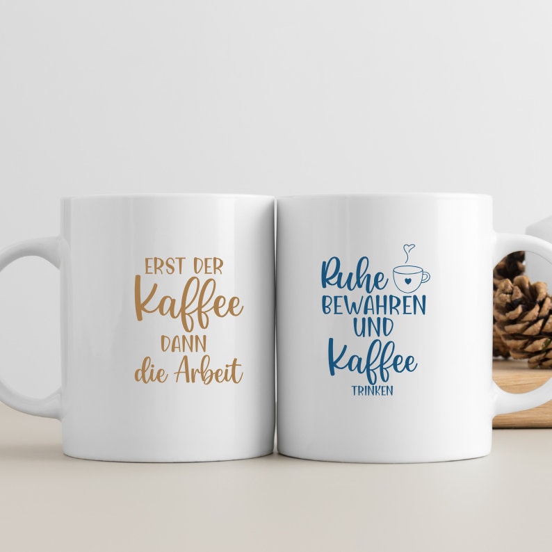 May include: Two white ceramic mugs with black handles. The mug on the left has the text "Erst der Kaffee dann die Arbeit" in brown lettering. The mug on the right has the text "Ruhe bewahren und Kaffee trinken" in blue lettering.