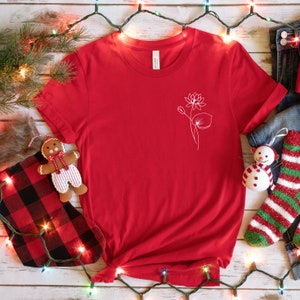 May include: Red t-shirt with a white outline of a flower and leaves on the front. The shirt is lying on a white wooden surface with Christmas lights and decorations.
