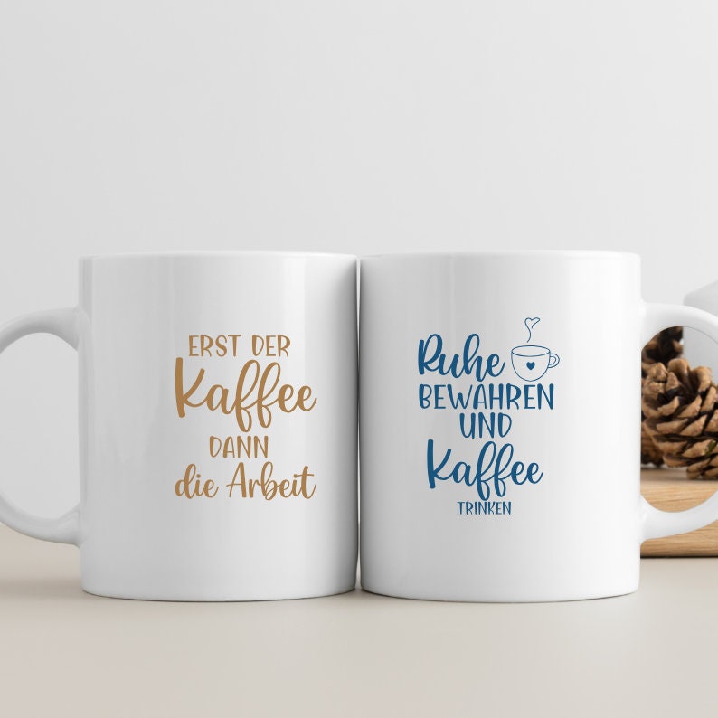 May include: Two white ceramic mugs with black handles. The mug on the left has the text "Erst der Kaffee dann die Arbeit" in brown lettering. The mug on the right has the text "Ruhe bewahren und Kaffee trinken" in blue lettering.