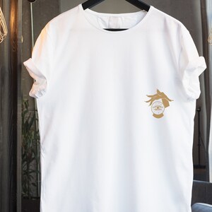 May include: White t-shirt with a gold graphic of a hand holding a crystal ball with an eye inside. The graphic is on the left side of the shirt.