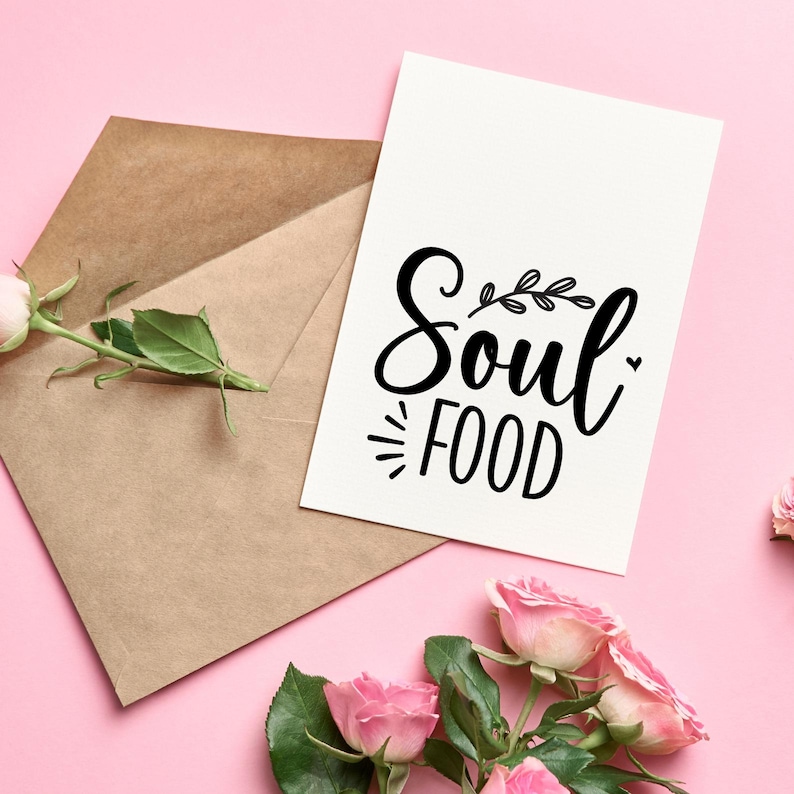 Op de afbeelding: Een witte kaart met de woorden "Soul Food" in zwart schrift en een bruine envelop liggen op een roze ondergrond. Roze rozen en groene bladeren zijn rond de kaart en envelop gerangschikt. De kaart heeft een decoratief bladerontwerp.