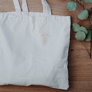 May include: White canvas tote bag with a gold embroidered design of a crescent moon and stars with the text "Moon & Co."