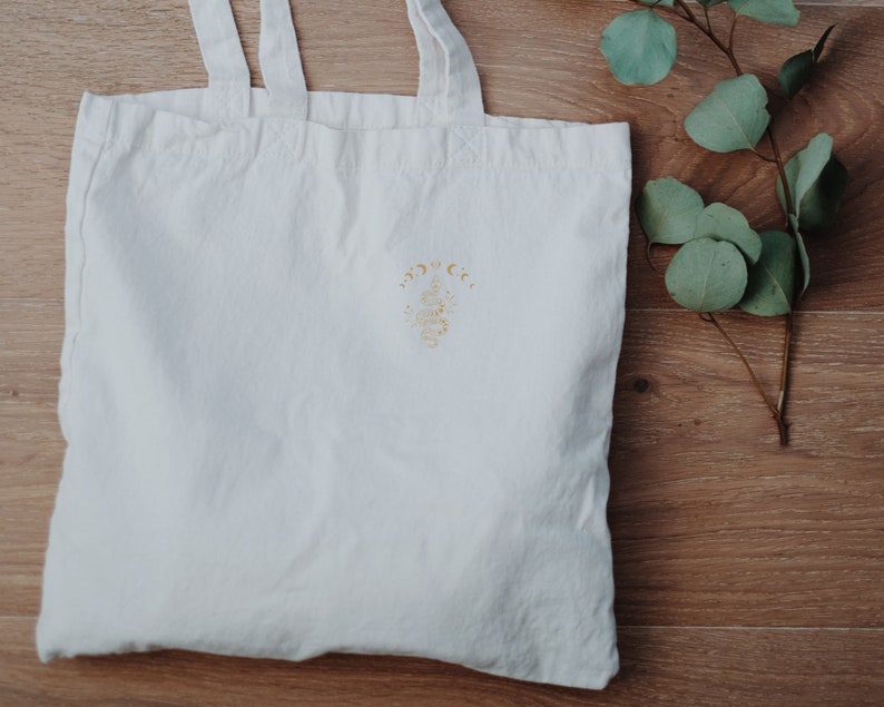 May include: White canvas tote bag with a gold embroidered design of a crescent moon and stars with the text "Moon & Co."