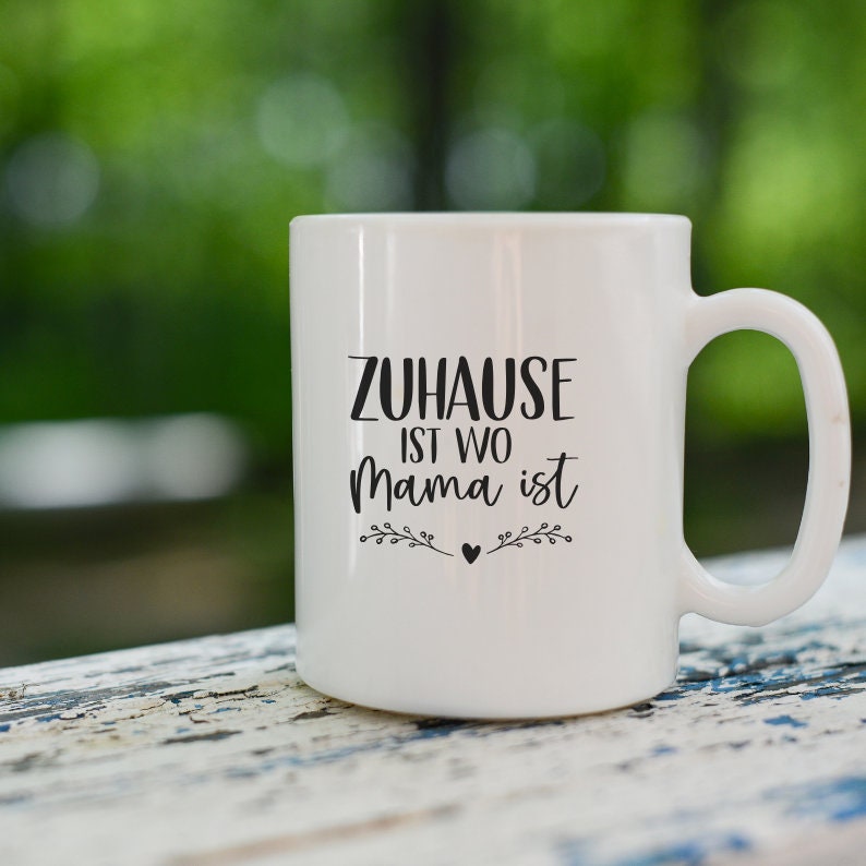 Pu&ograve; includere: Tazza in ceramica bianca con un designo nero che recita "Zuhause ist wo Mama ist" con un piccolo cuore e dettagli floreali.