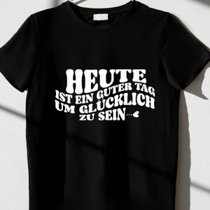 Peut inclure: T-shirt noir avec un texte blanc qui dit "HEUTE IST EIN GUTER TAG UM GL&Uuml;CKLICH ZU SEIN..." avec un petit symbole de c&oelig;ur.