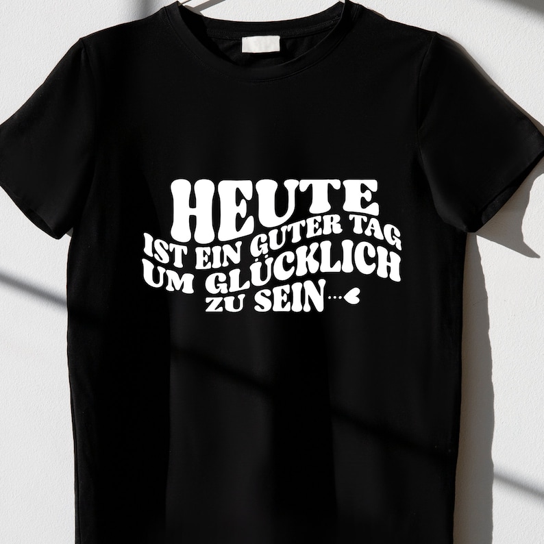 Peut inclure: T-shirt noir avec un texte blanc qui dit "HEUTE IST EIN GUTER TAG UM GL&Uuml;CKLICH ZU SEIN..." avec un petit symbole de c&oelig;ur.