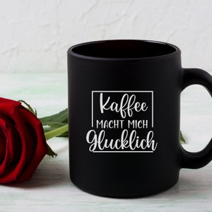 May include: Black coffee mug with white text that reads "Kaffee macht mich glucklich" which translates to "Coffee makes me happy".