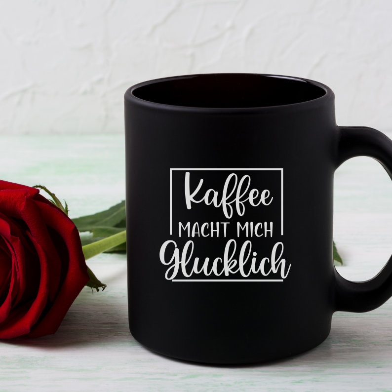 May include: Black coffee mug with white text that reads "Kaffee macht mich glucklich" which translates to "Coffee makes me happy".