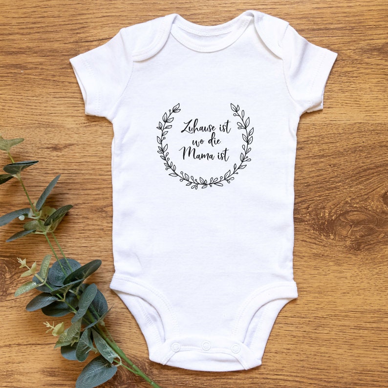 Pu&ograve; includere: Body bianco per neonato con un design a corona nera e il testo "Zuhause ist wo die Mama ist" in un carattere nero.
