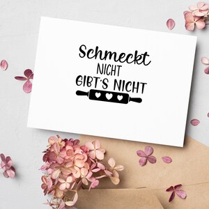 May include: A white card with black text that reads "Schmeckt nicht gibt's nicht" with a rolling pin and three hearts underneath. The card is on a brown surface with pink flowers.