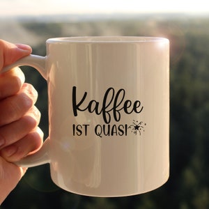 May include: White ceramic coffee mug with black text that reads "Kaffee ist quasi" with a small firework graphic.