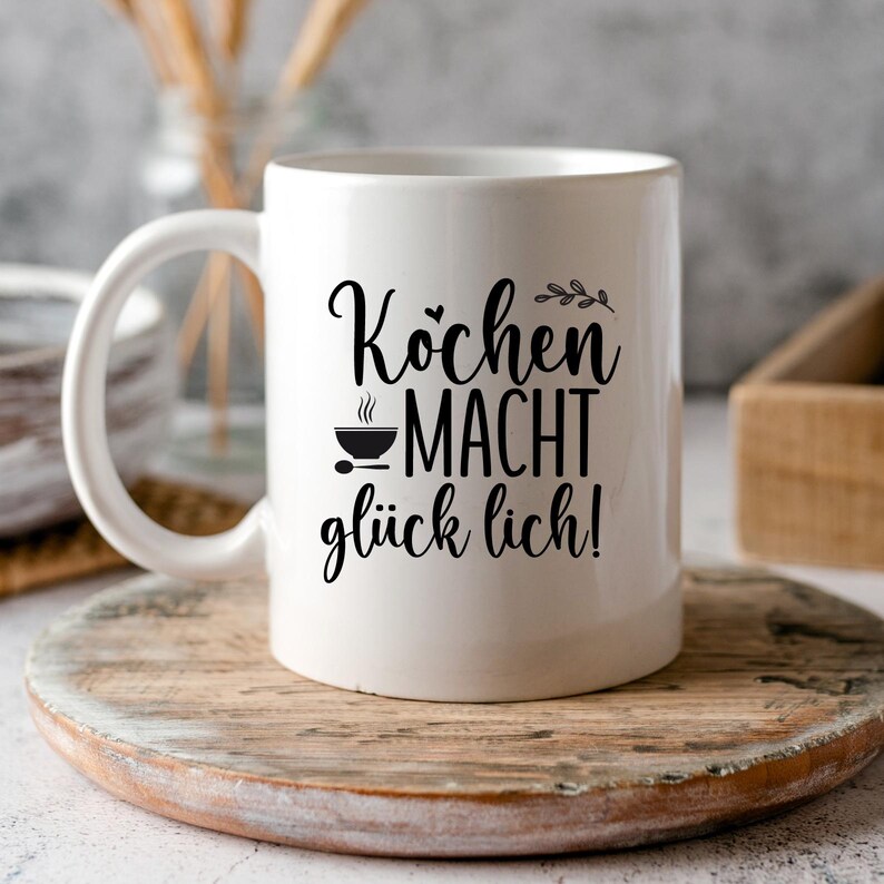 Op de afbeelding: Witte keramische mok met de Duitse tekst "Kochen macht gl&uuml;cklich!" in zwart schrift. De mok heeft een handvat en een afbeelding van een kom met stoom en een lepel. De mok staat op een houten onderzetter.