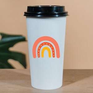 Puede incluir: Una taza de caf&eacute; de papel blanco con tapa negra. La taza presenta un dise&ntilde;o de arco&iacute;ris colorido en tonos naranja y amarillo, con estrellas blancas. La taza est&aacute; sobre una superficie de madera.