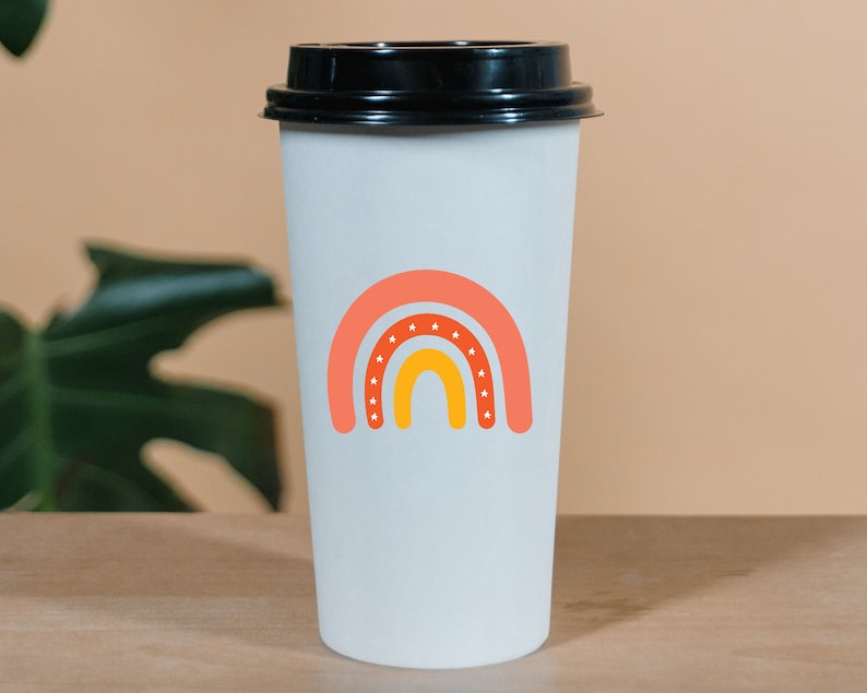 Puede incluir: Una taza de caf&eacute; de papel blanco con tapa negra. La taza presenta un dise&ntilde;o de arco&iacute;ris colorido en tonos naranja y amarillo, con estrellas blancas. La taza est&aacute; sobre una superficie de madera.