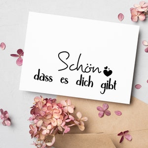 May include: White card with black text that reads "Sch&ouml;n dass es dich gibt" with a small black heart symbol. The card is surrounded by pink flowers.
