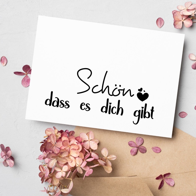 May include: White card with black text that reads "Sch&ouml;n dass es dich gibt" with a small black heart symbol. The card is surrounded by pink flowers.