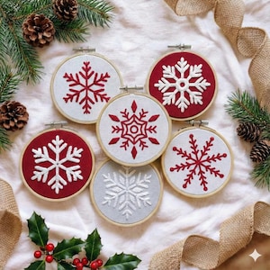 May include: Six embroidered snowflake ornaments in wooden hoops. The snowflakes are red and white, set against red and white fabric backgrounds. The ornaments are arranged with pine needles, pine cones, and burlap ribbon.