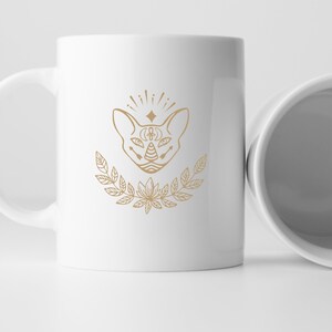 May include: White ceramic mug with a gold outline of a cat's face with a third eye and a floral design.