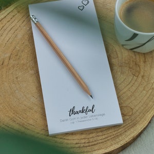 Könnte beinhalten: Ein weißer Notizblock mit einem Bleistift darauf. Der Notizblock hat oben zwei kleine Herzen gezeichnet und die Worte "thankful" und "Dankt Gott in jeder Lebenslage (vgl. 1. Thessalonicher 5,18)" darauf gedruckt.