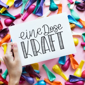 Könnte beinhalten: Eine weiße Karte mit schwarzer Schrift, auf der "eine Dose Kraft" steht, umgeben von bunten Luftballons. Die Karte wird von einer Hand gehalten.