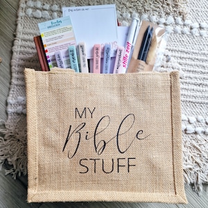 Könnte beinhalten: Eine Jutetasche mit der Aufschrift "MY Bible STUFF", gefüllt mit Schreibwaren. Enthalten sind farbige Lesezeichen, Stifte, Bleistifte und ein kleines Notizbuch. Die Tasche steht auf einem strukturierten, neutralfarbenen Teppich.