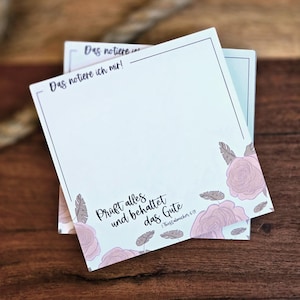 Könnte beinhalten: Ein Stapel hellblauer Notizblöcke mit einem floralen Design mit rosa Rosen und braunen Blättern. Der Notizblock hat einen schwarzen Rand und den Text "Das notiere ich mit!" und "Prüft alles und behaltet das Gute 1 Thessalonicher 5:18" darauf gedruckt.