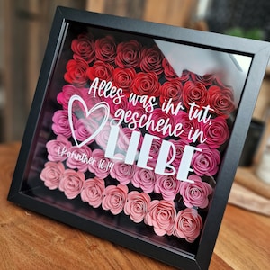 Könnte beinhalten: Gerahmtes Kunstwerk mit Papierrosen in Rot- und Rosatönen. Der weiße Text lautet "Alles was ihr tut, geschehe in LIEBE" und "1.Korinther 16.14". Der schwarze Rahmen umschließt die Rosen, die hinter Glas präsentiert werden.