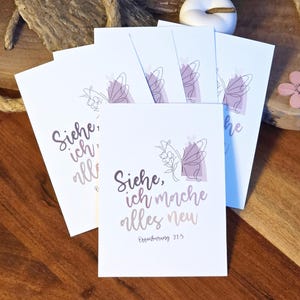 Könnte beinhalten: Eine Reihe weißer Karten mit einem Schmetterlingsdesign und dem Text "Siehe, ich mache alles neu". Der Schmetterling ist lila und der Text in Lila- und Brauntönen gehalten. Die Karten liegen auf einer Holzoberfläche.