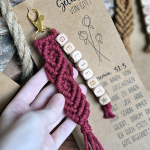 Könnte beinhalten: Ein bordeauxfarbener Makramee-Schlüsselanhänger mit einem goldfarbenen Verschluss. Holzbuchstabenwürfel schreiben "GELIEBT". Der Hintergrund zeigt eine Karte mit deutschem Text und floralen Illustrationen.