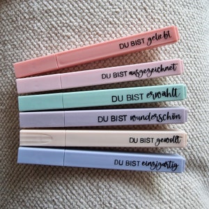 Könnte beinhalten: Sechs Textmarker in Pastellfarben, darunter Rosa, Lila, Mint, Lavendel, Creme und Blau. Jeder Textmarker trägt einen deutschen Satz in schwarzer Schrift. Die Textmarker liegen auf einer strukturierten, neutralfarbenen Oberfläche.