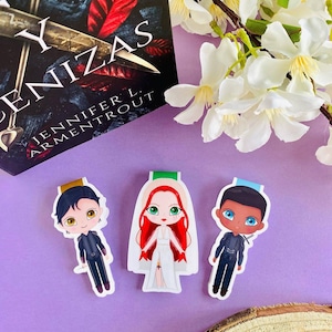 May include: Three character bookmarks featuring illustrations of characters from the book "From Blood and Ash" by Jennifer L. Armentrout. The bookmarks are printed on white cardstock and feature a black-haired male character, a red-haired female character, and a brown-skinned male character.