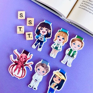 Puede incluir: Seis marcadores de colores con personajes de dibujos animados y un monstruo rojo con múltiples patas. Los marcadores están sobre una superficie morada con fichas de letras de madera que deletrean "S, T, S, T".