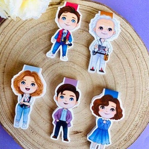 May include: Five colorful character bookmarks on a wooden surface. The bookmarks feature cartoon illustrations of characters in various outfits. The characters have different hair colors and are wearing blue, red, purple, and white outfits. The bookmarks are on a wooden surface.