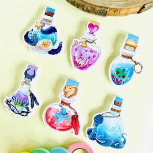 May include: Six colorful glass bottle bookmarks with different designs. Each bottle has a cork stopper and a ribbon attached. The designs include an octopus, a heart, a skull, a genie, a mermaid, and a feather.