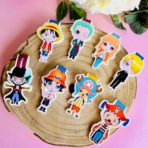 Puede incluir: Colección de marcapáginas de personajes de anime coloridos, con varios diseños sobre una rodaja de madera. Los personajes tienen atuendos y expresiones distintivas, con colores vibrantes y detalles. Cada marcapáginas tiene una pequeña pestaña en la parte superior.