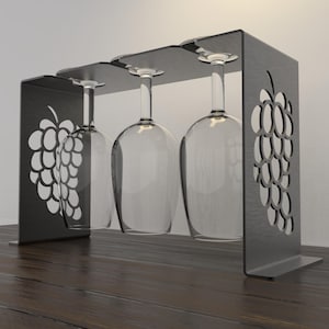 May include: A dark gray metal wine glass rack with a grape design cut out on the sides. The rack holds three clear wine glasses. The rack is sitting on a dark wooden surface.
