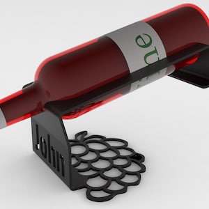 Peut inclure: Un porte-bouteille de vin en métal noir avec un motif de grappe de raisin découpé sur la base et le nom John découpé au laser sur le support vertical. Le support est incliné pour maintenir une bouteille en verre remplie de liquide rouge.