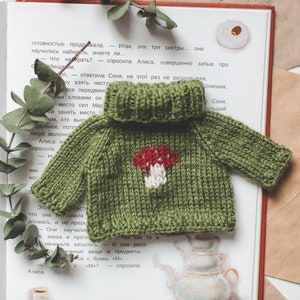 May include: A green knitted jumper with a white mushroom design. The jumper is lying on a book with text in Russian.