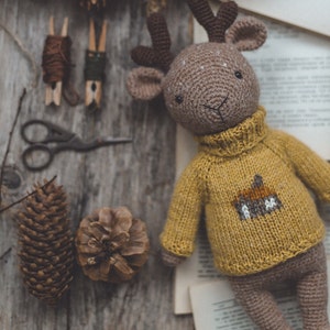 May include: A crocheted deer toy wearing a yellow jumper with a brown deer design. The toy is lying on a wooden surface with pine cones and other craft supplies.