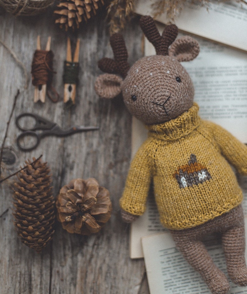 May include: A crocheted deer toy wearing a yellow jumper with a brown deer design. The toy is lying on a wooden surface with pine cones and other craft supplies.