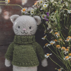 May include: A crocheted cat toy wearing a green sweater, with white fur. The cat is positioned in front of a bunch of white daisies.