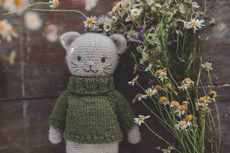 May include: A crocheted cat toy wearing a green sweater, with white fur. The cat is positioned in front of a bunch of white daisies.