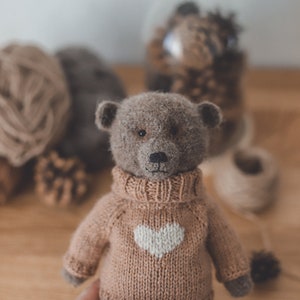 May include: A brown teddy bear wearing a knitted jumper with a white heart design. The jumper is a light brown colour. The bear is sitting on a wooden surface.