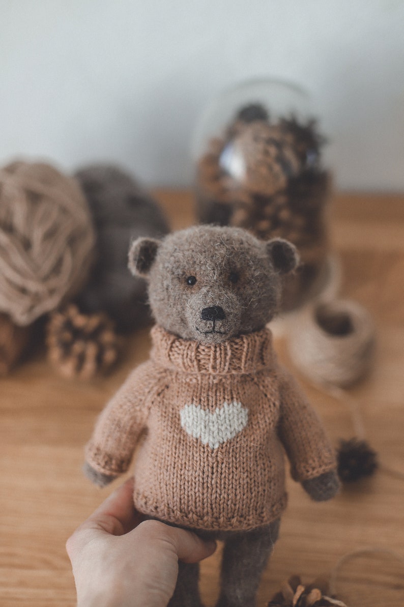 May include: A brown teddy bear wearing a knitted jumper with a white heart design. The jumper is a light brown colour. The bear is sitting on a wooden surface.