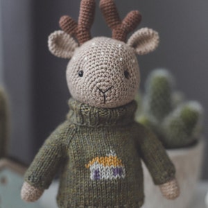 May include: A crocheted deer toy wearing a green knitted jumper with a small house design on the front. The deer has brown antlers and a beige body.