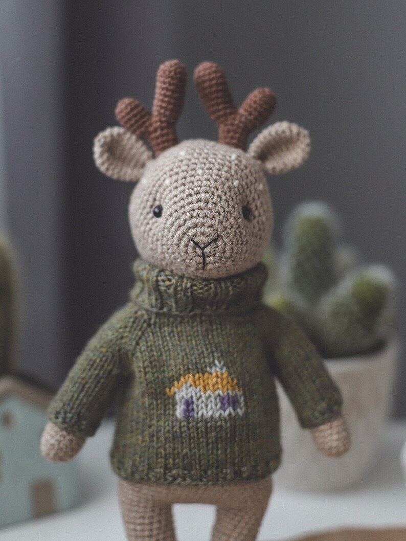 May include: A crocheted deer toy wearing a green knitted jumper with a small house design on the front. The deer has brown antlers and a beige body.