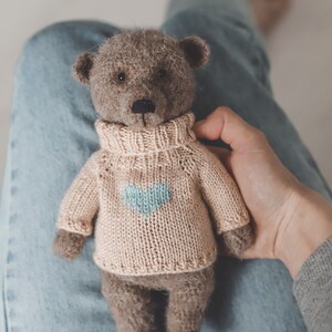 May include: A brown teddy bear wearing a light pink knitted jumper with a light blue heart design. The bear is sitting on someone's lap.