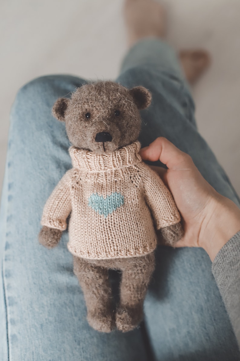 May include: A brown teddy bear wearing a light pink knitted jumper with a light blue heart design. The bear is sitting on someone's lap.