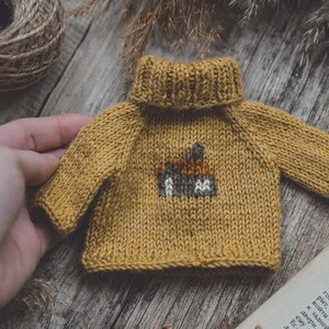 May include: A small, mustard yellow knitted jumper with a brown and white embroidered design on the front. The jumper has a turtleneck.